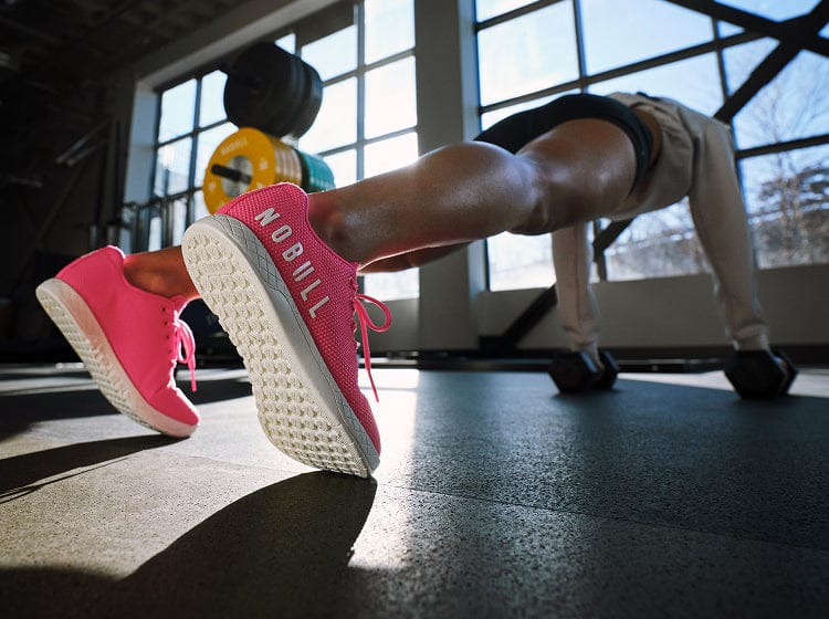 Person exercising with pink 'NOBULL' sneakers on a gym floor.