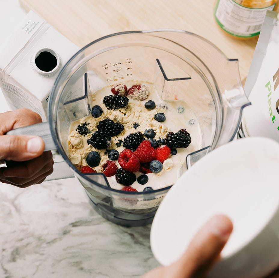 Blender with ingredients including plant protein, berries and a hand holding the lid on a marble countertop.