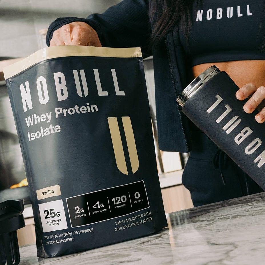 Person holding NOBULL whey protein isolate package and tumbler in a kitchen setting.