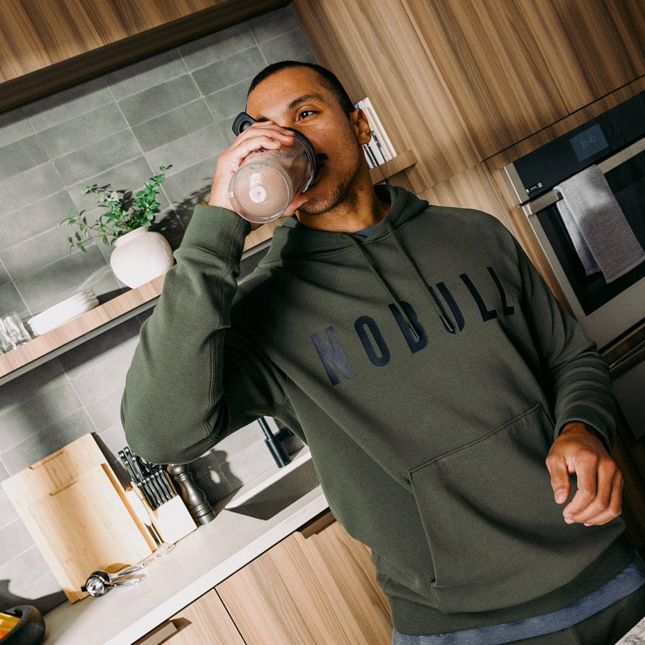 Man drinking from a glass in a kitchen wearing a hoodie with 'NOULL' branding.
