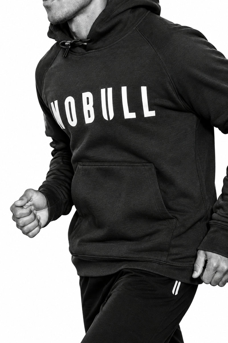 A person wearing a muted sage green hoodie with the word 'NOBULL' printed in white across the chest, shown in a dark indoor setting.