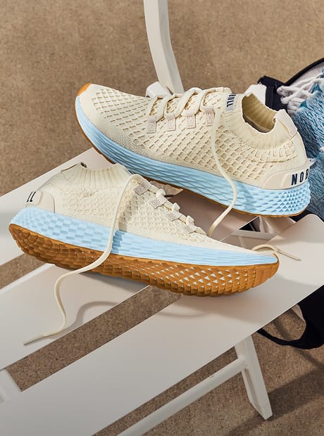 A pair of knit athletic sneakers resting on a white bench. The shoes feature a cream-colored knit upper, a light blue midsole, and a brown textured outsole.