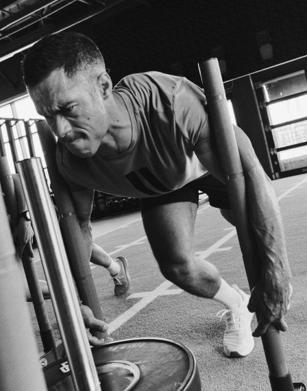 A black and white action photograph of a male athlete in a gym, leaning forward and gripping a vertical training pole. He is wearing a short-sleeve athletic shirt, shorts, and athletic shoes on a track field surface.