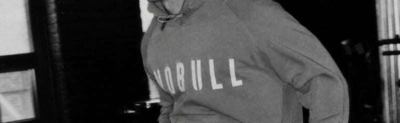 A person wearing a muted sage green pullover hoodie with the word 'NOBULL' printed in large white letters across the chest, shown in a dim indoor setting.