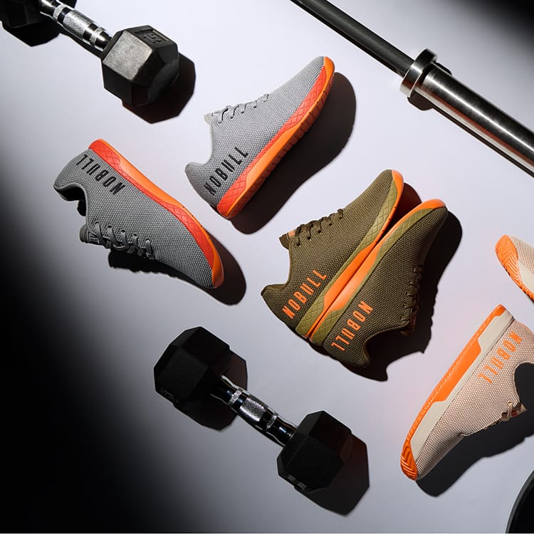 Product image. A styled flat-lay of several athletic sneakers arranged on a light surface with dumbbells and a metal bar as props. The shoes show multiple colorways: gray with orange, olive green with orange, and beige with orange.