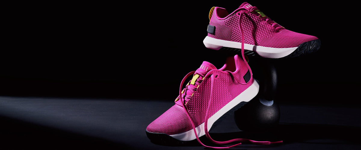 A pair of bright pink NOBULL Product athletic sneakers displayed on a black studio stand, with one shoe resting on the surface and the other elevated above it.