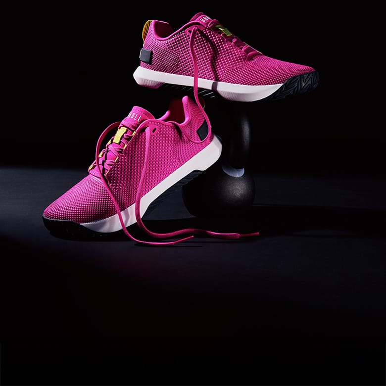 A pair of bright pink NOBULL Product athletic sneakers displayed on a black studio stand, with one shoe resting on the surface and the other elevated above it.