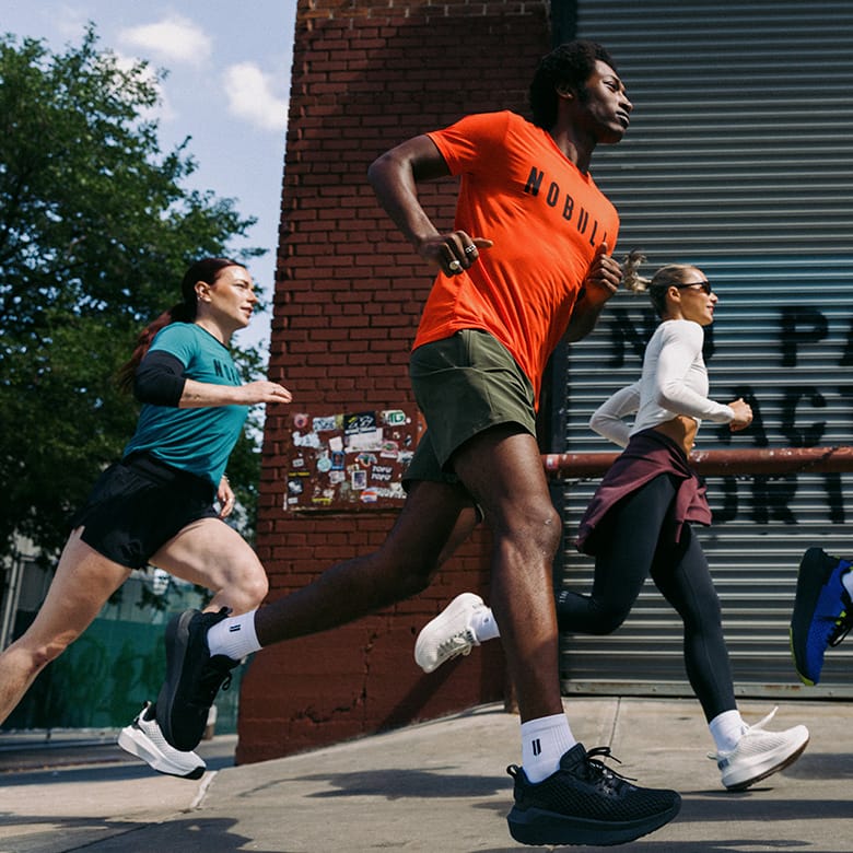 Product on Body. Three runners jogging outdoors beside a brick wall and metal garage door. The central runner wears a bright orange t-shirt with NOBULL' printed on the front, olive green shorts, white socks, and black running shoes.