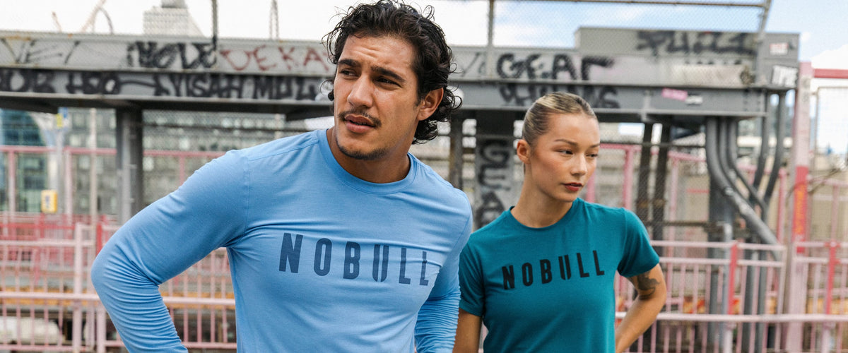 Promo - Product on body. A man and a woman wearing NOBULL athletic t-shirts in a construction site setting. The man in the foreground wears a light blue long-sleeve t-shirt with 'NOBULL' printed across the chest, while the woman behind him wears a teal-blue short-sleeve t-shirt with 'NOBULL' printed on the front.
