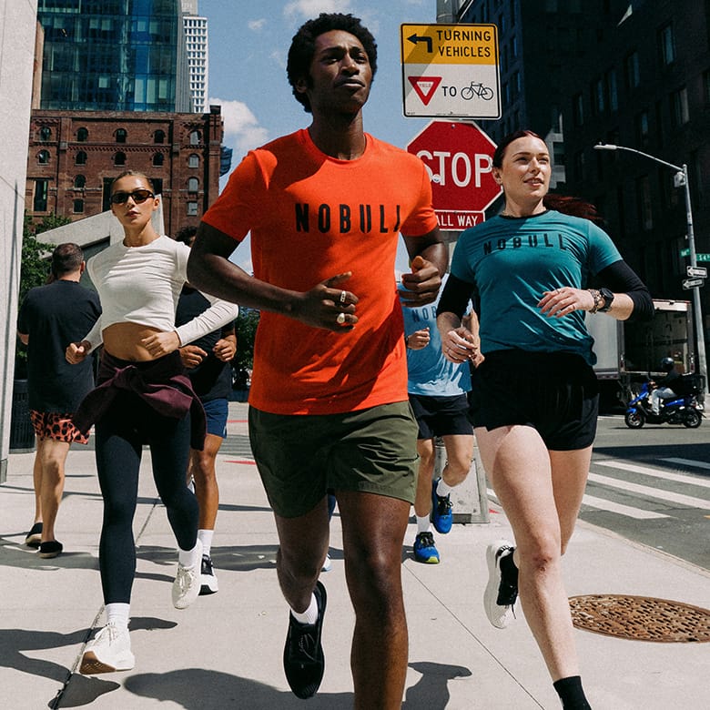 Model jogging on a city street, with the central figure wearing a bright orange t-shirt with the word 'NOBULL' printed across the chest. Other runners in teal and white are visible behind them, surrounded by urban buildings and street signs.