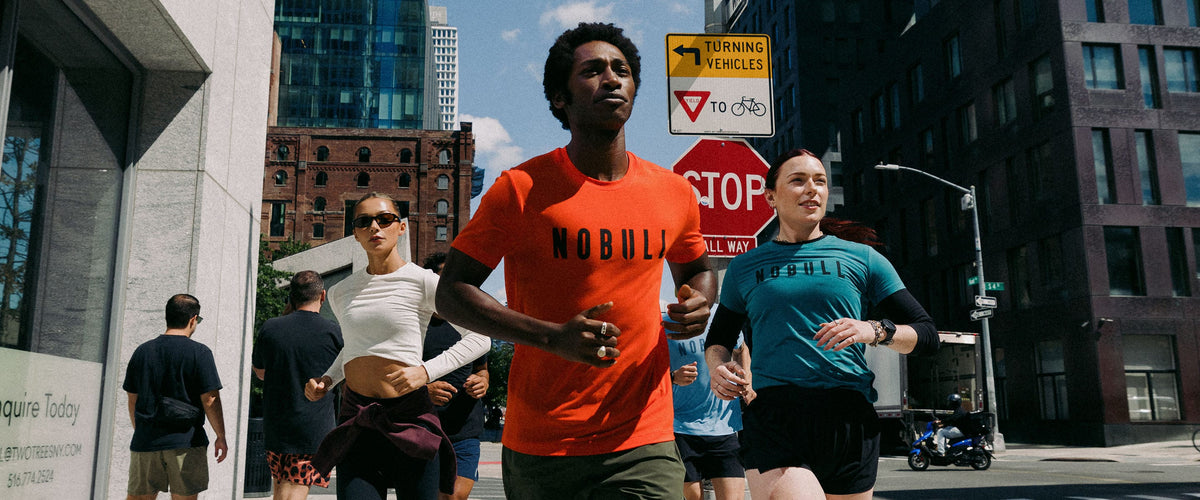 Model jogging on a city street, with the central figure wearing a bright orange t-shirt with the word 'NOBULL' printed across the chest. Other runners in teal and white are visible behind them, surrounded by urban buildings and street signs.
