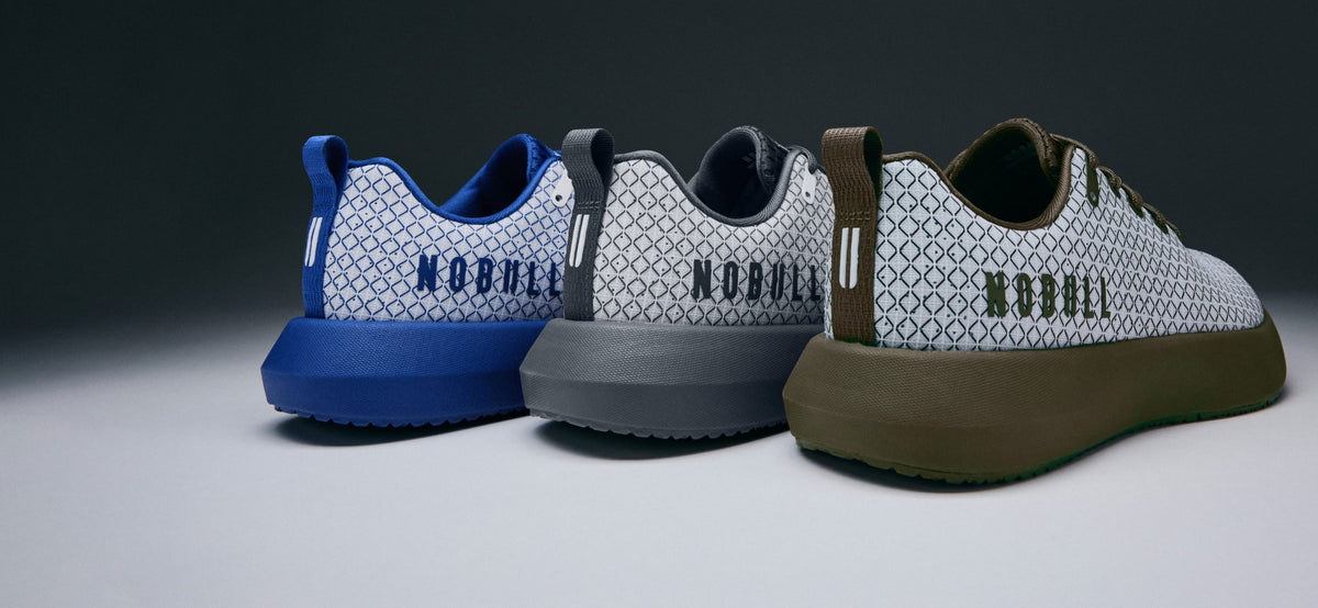 product - Three low-top sneakers from NOBULL are lined up on a dark-to-light gradient studio background. The sneakers feature a white geometric patterned upper, a thick blue, gray, and olive-brown sole, and the brand name 'NOBULL' printed on the heel.