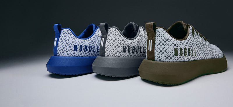 product - Three low-top sneakers from NOBULL are lined up on a dark-to-light gradient studio background. The sneakers feature a white geometric patterned upper, a thick blue, gray, and olive-brown sole, and the brand name 'NOBULL' printed on the heel.