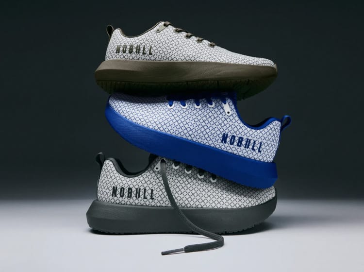 product - Three low-top sneakers stacked in a studio setup against a dark gray background. The shoes have a white and gray geometric patterned upper, with one featuring olive green soles, one with a bright blue sole, and one with dark gray soles.