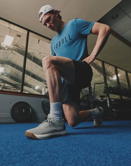 A man in a gym wearing a blue t-shirt with 'KOBULL' printed on the chest, black shorts, white socks, and gray running shoes, performing a lunge on a blue gym floor.