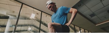 A man in a blue t-shirt and white cap climbing a glass wall in a modern indoor setting.
