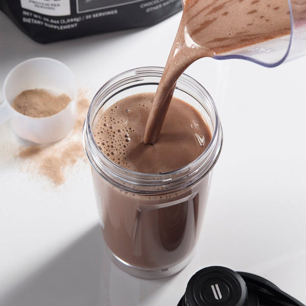 Chocolate powder being poured into a glass jar on a white surface.