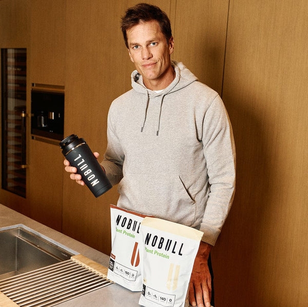 Tom Brady holding the NOBULL shaker bottle with plant protein packages